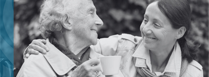 A Long Term Care Resident and a Volunteer are smiling at each other while the resident is drinking a cup of tea.