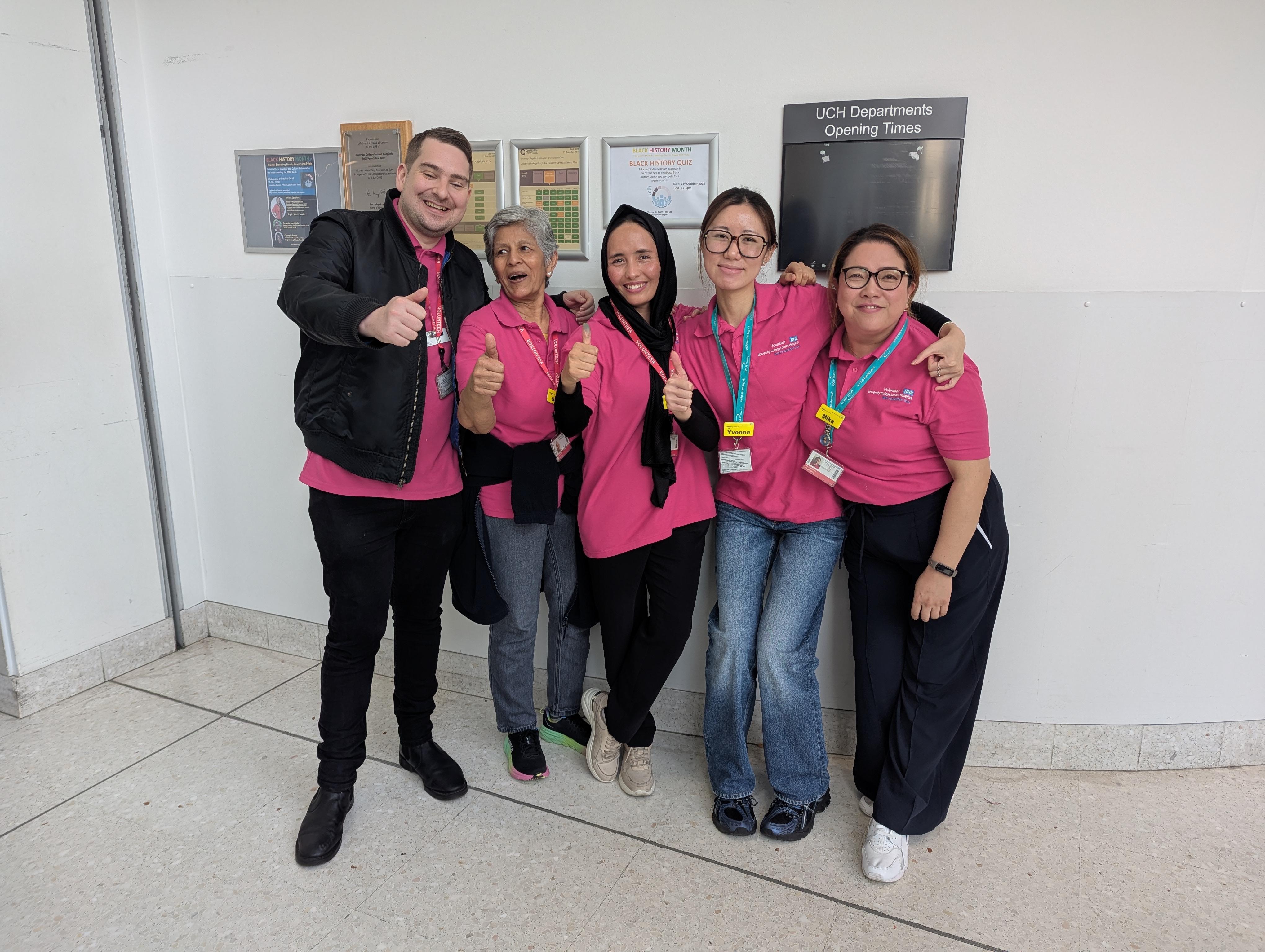 Five hospital volunteers wearing bright pink polo shirts stand together smiling with their arms around one another in a hospital corridor, giving thumbs-up gestures.