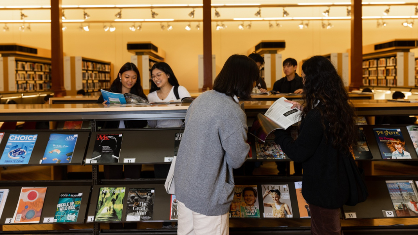 Two people stand with their backs to the viewer. They are looking at books displayed in the Library. Other Library visitors can be seen in the background. 